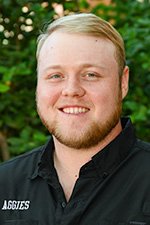Greg Brooks headshot. He is wearing a black shirt with the word Aggies written on the right chest. He is standing in front of green foliage.