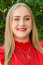 Hadley Tokraks headshot. She is wearing a red shirt with jewelry in front of green foliage. 