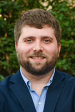 Image of Dr. William Norris, Assistant Professor in the Department of Agricultural and Extension Education at NMSU. He has brown hair with a beard, and is wearing a grey and black polo shirt while standing in front of a grey background.