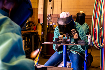 Image of a student welding in the agricultural education shop class. Student is wearing a face shield, teal protective coat, and heavy welding gloves.